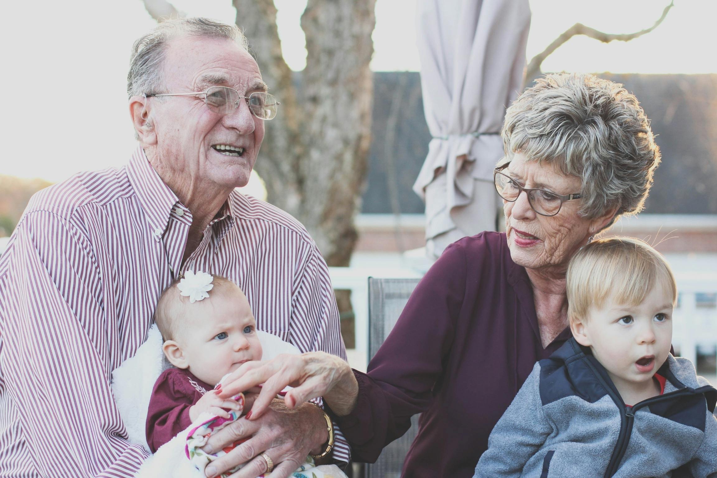 Grandparents in their 70s sit side by side, each with a grandbaby in their lap.